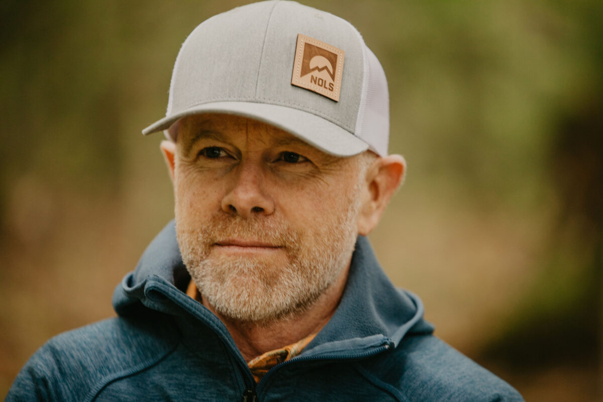 a man with a grey NOLS baseball hat looks to the left of camera with a hint of a smile
