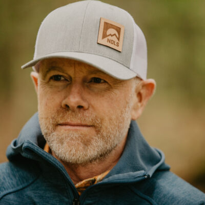 a man with a grey NOLS baseball hat looks to the left of camera with a hint of a smile