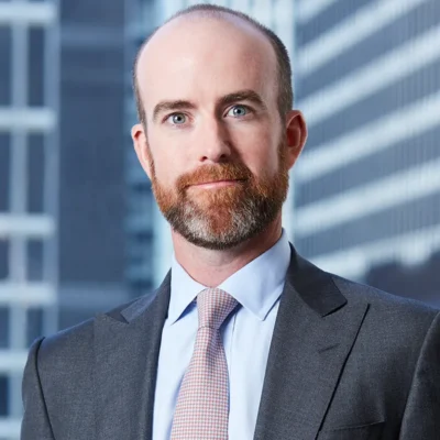 A man with a suit jacket and tie stands against a skyscraper background. He has a red mustache and beard.