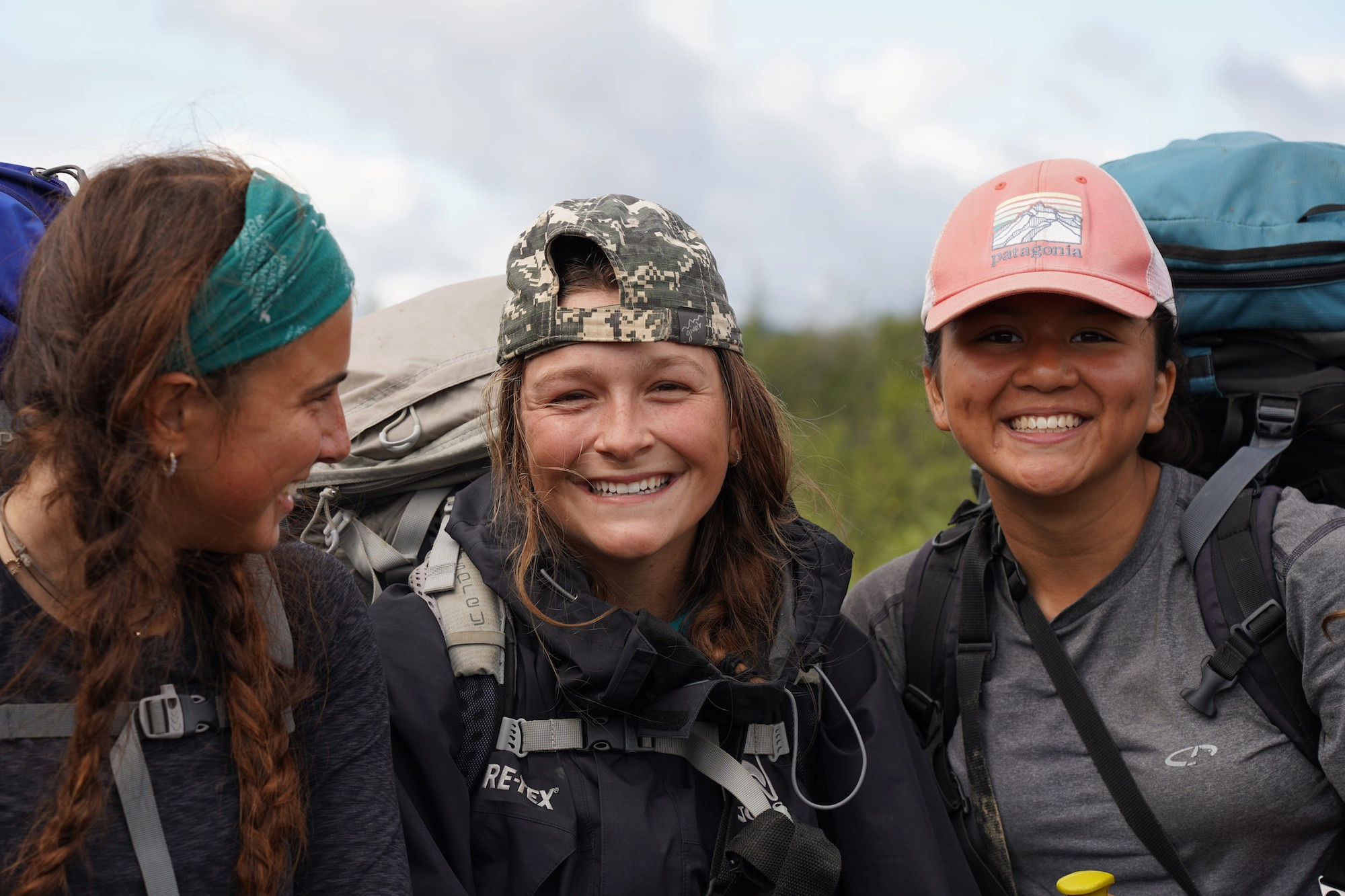 Three teen girls smile and pose for a photo. They have hiking backpacks and are dressed for the outdoors.