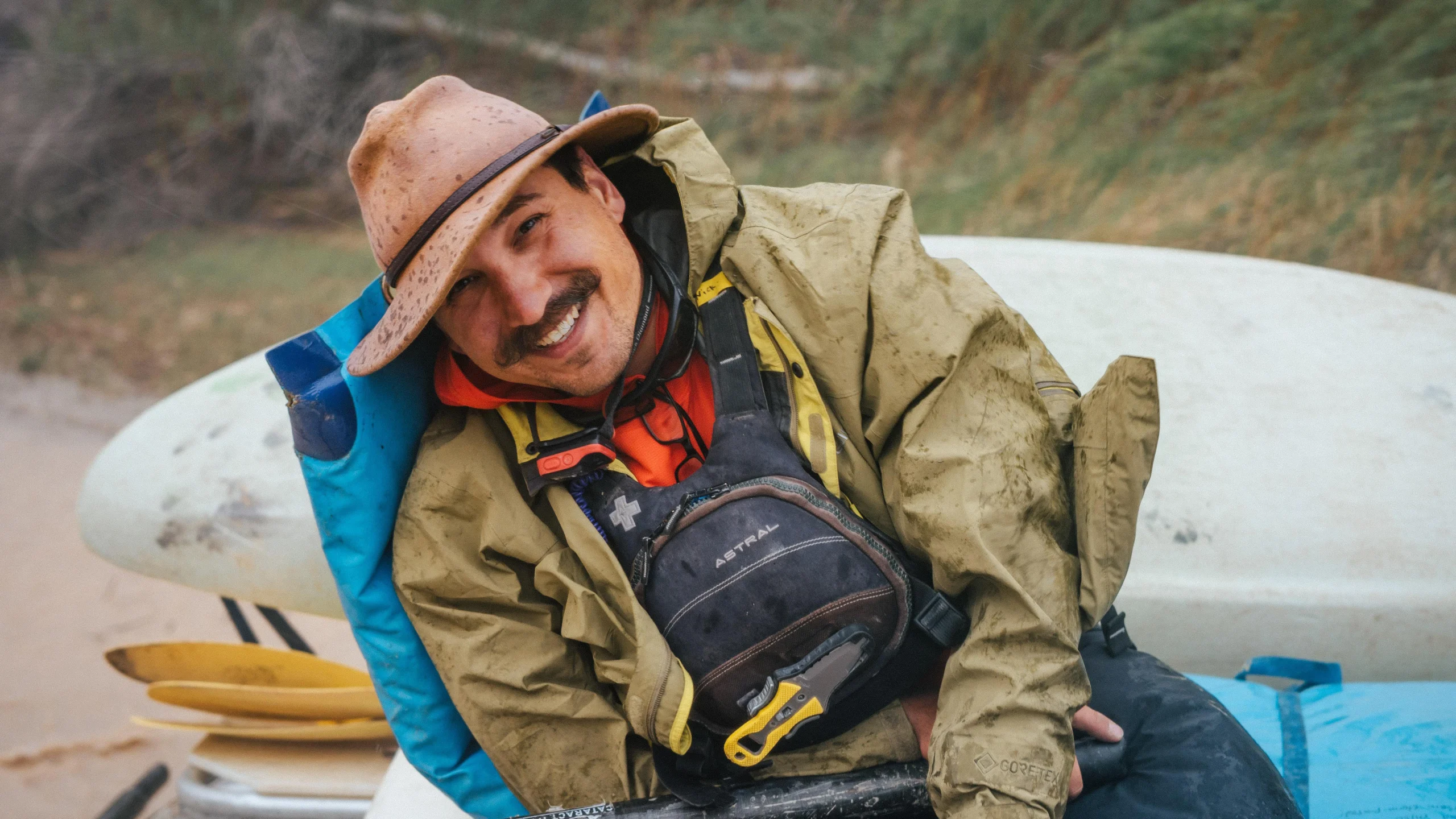 a man in a brimmed hat smiles at the camera while posed on a whitewater raft