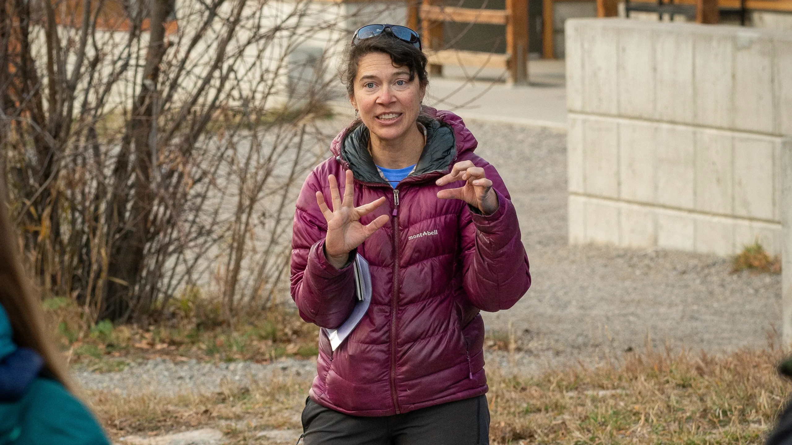 a woman in an eggplant-colored puffy jacket demonstrates something with her hands