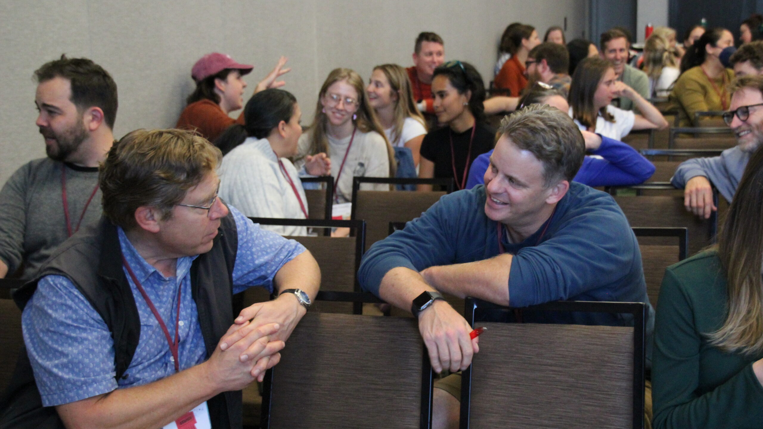 two men in a classroom talk to one another across a chair. many people in the background are engaged in conversation