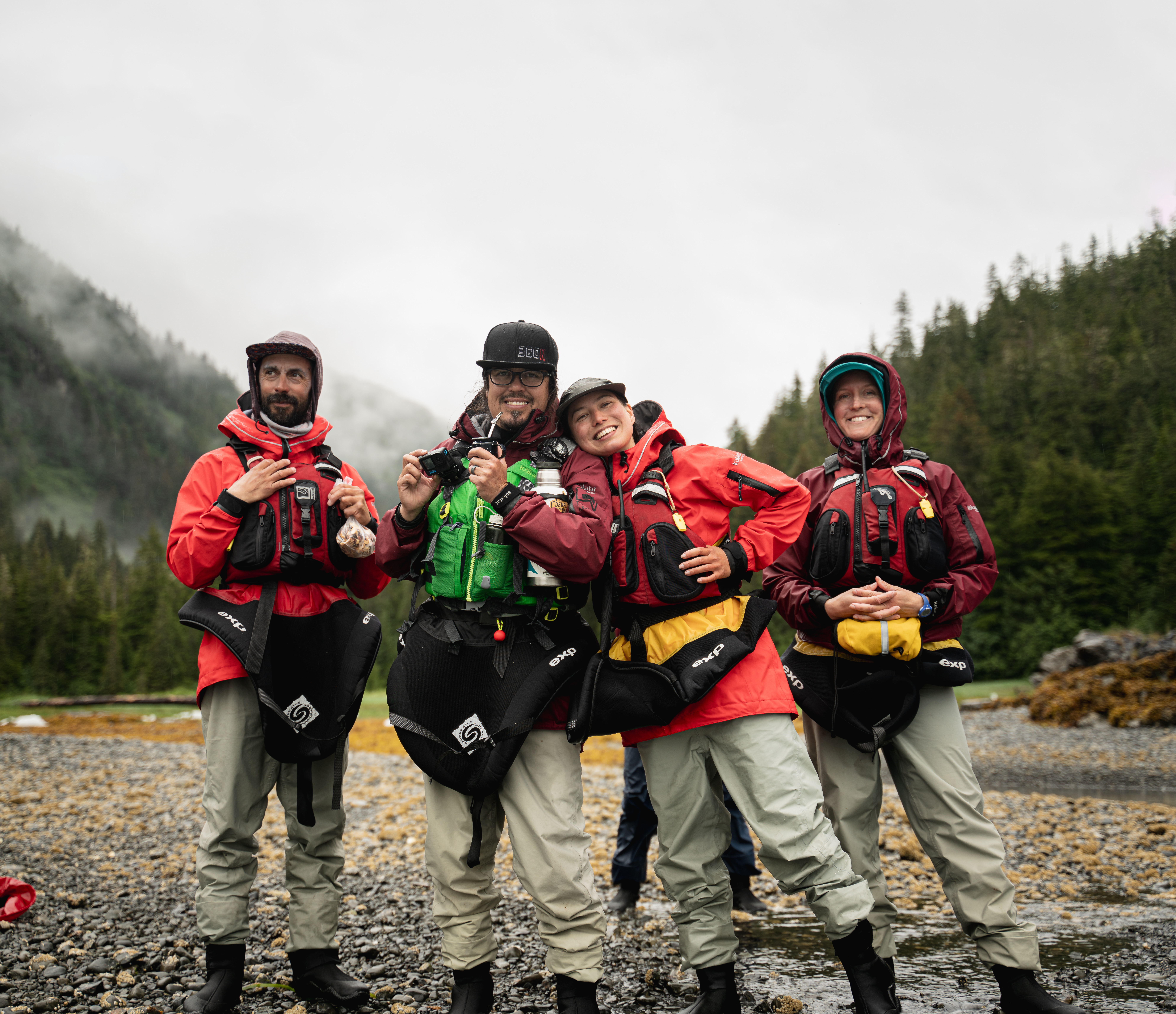 four people in sea kayak outfits pose on a cobble beach. the sky behind them is grey