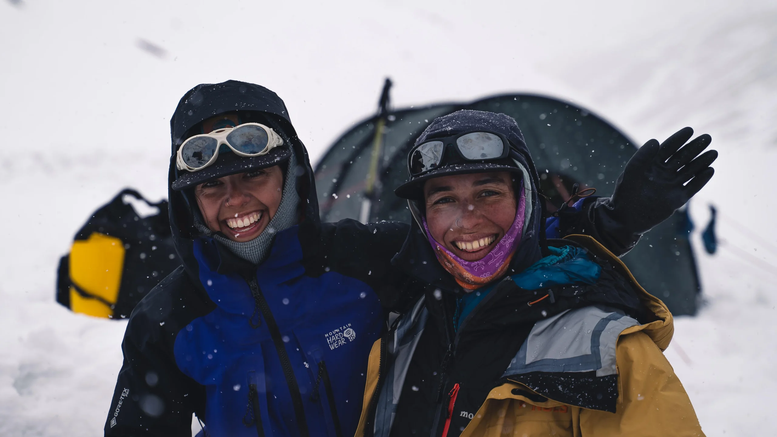 two women in cold weather clothing and sunglasses smile in a snowstorm