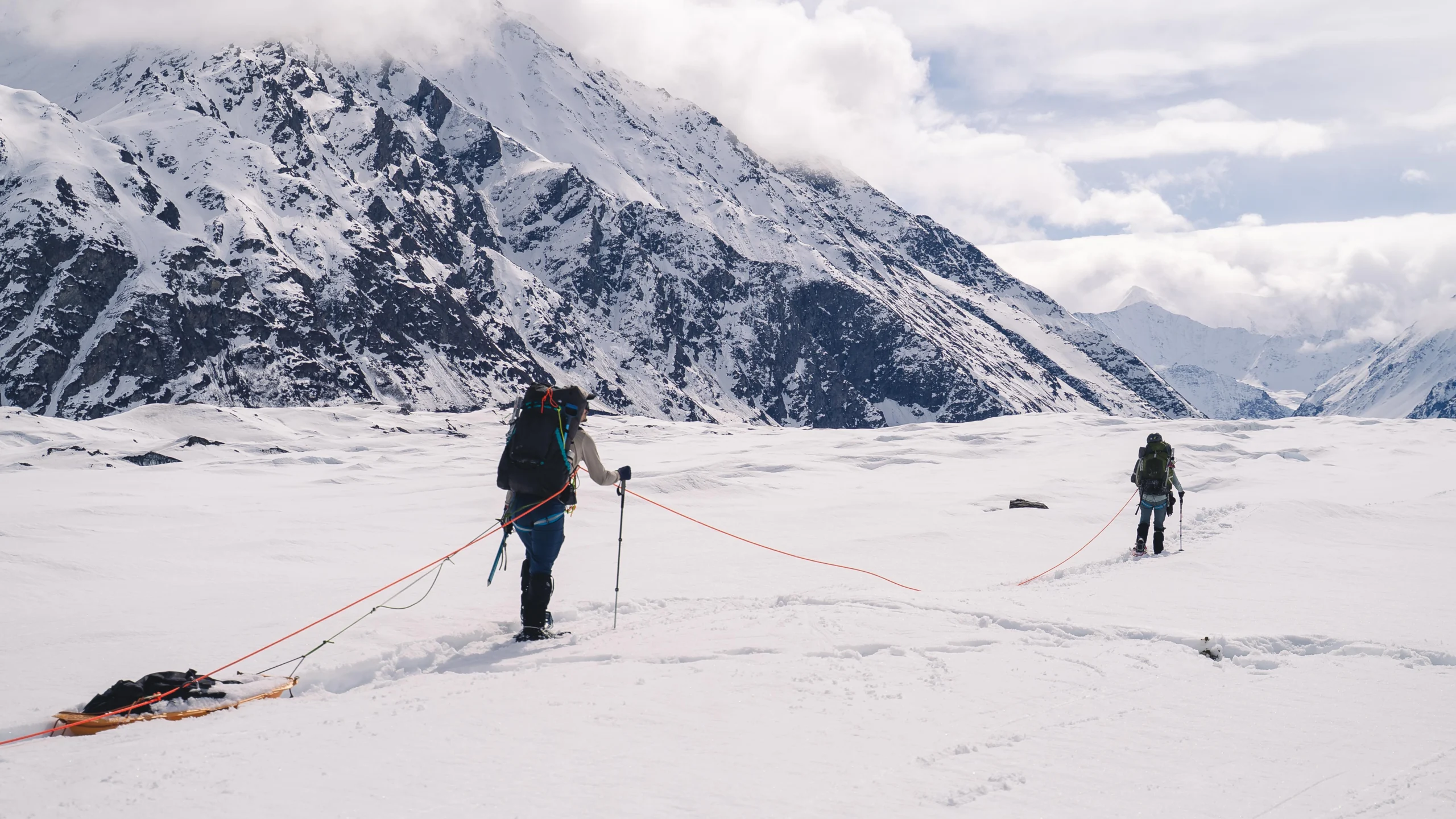 two people on a rope team haul sleds across a snowy expanse. there are mountains in the background