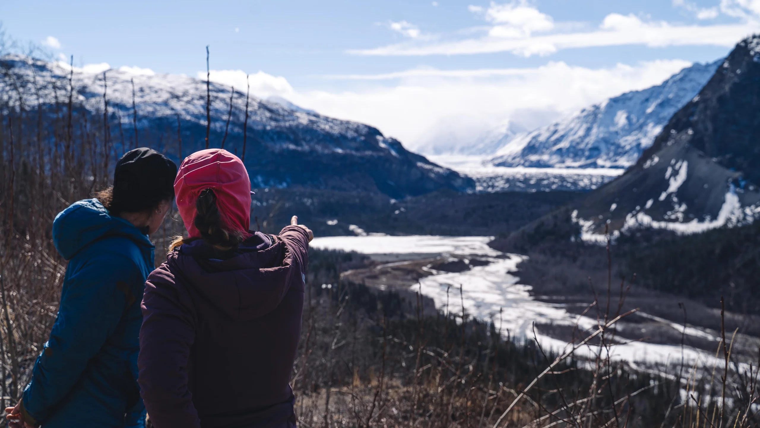two people in outdoor gear point out towards a glacial river and snowy mountains