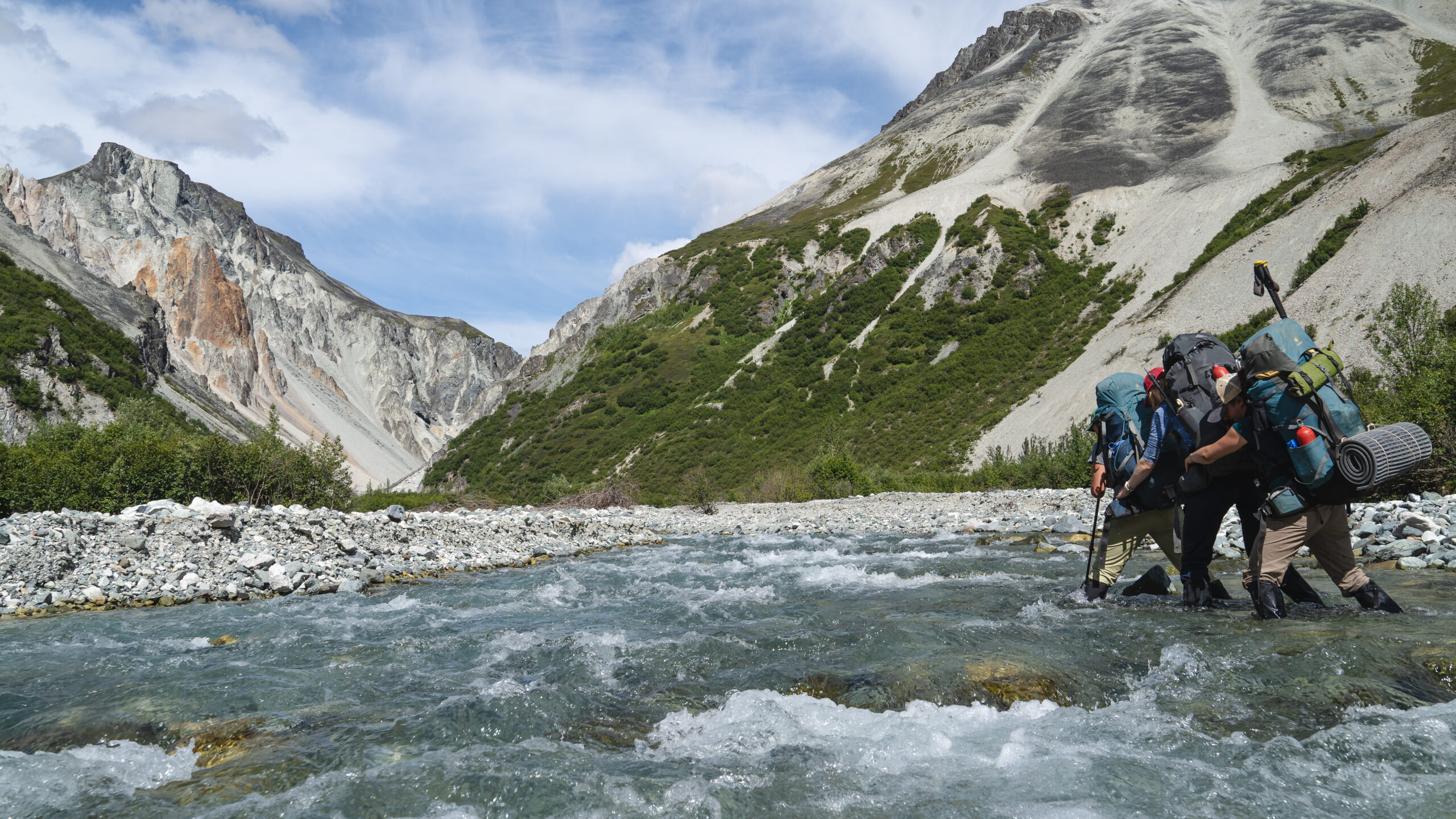 four people wearing large backpacks wade across a shallow stream in a line. there are mountains in the background.