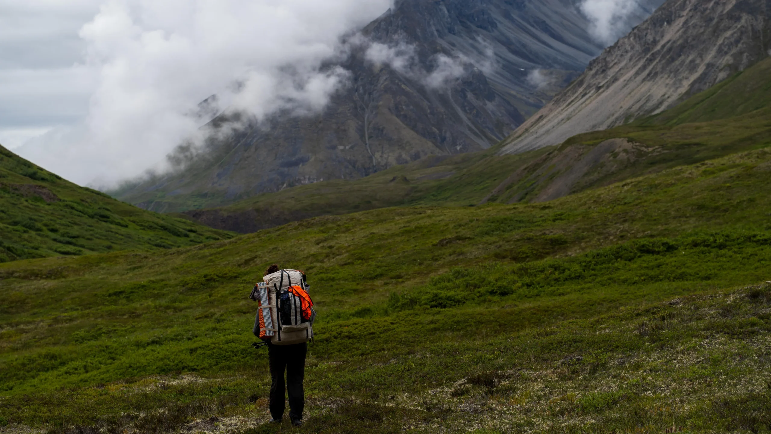 a person wearing a backpack stands in a valley looking out at large mountains blanketed in fog.