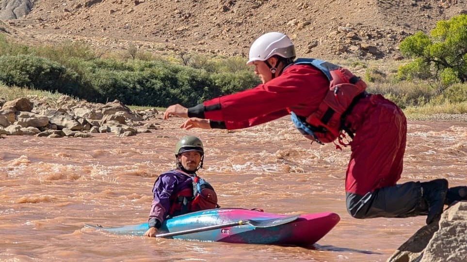 a man in a red drysuit and white helmet jumps into a muddy river. a person in a kayak watches him carefully