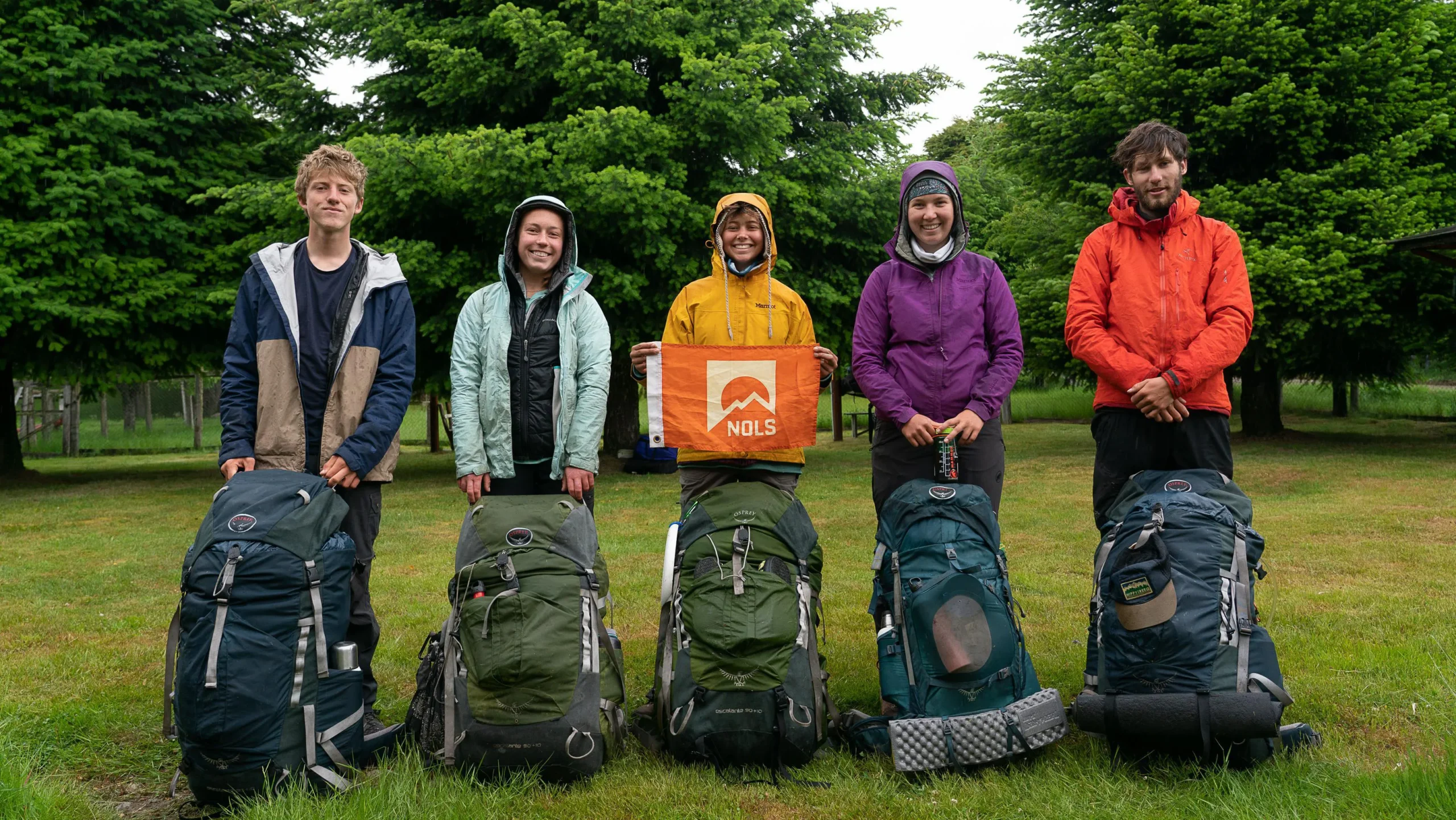 five people in outdoor gear post in a row with backpacks in front of their legs. the middle person is holding a flag with the NOLS logo