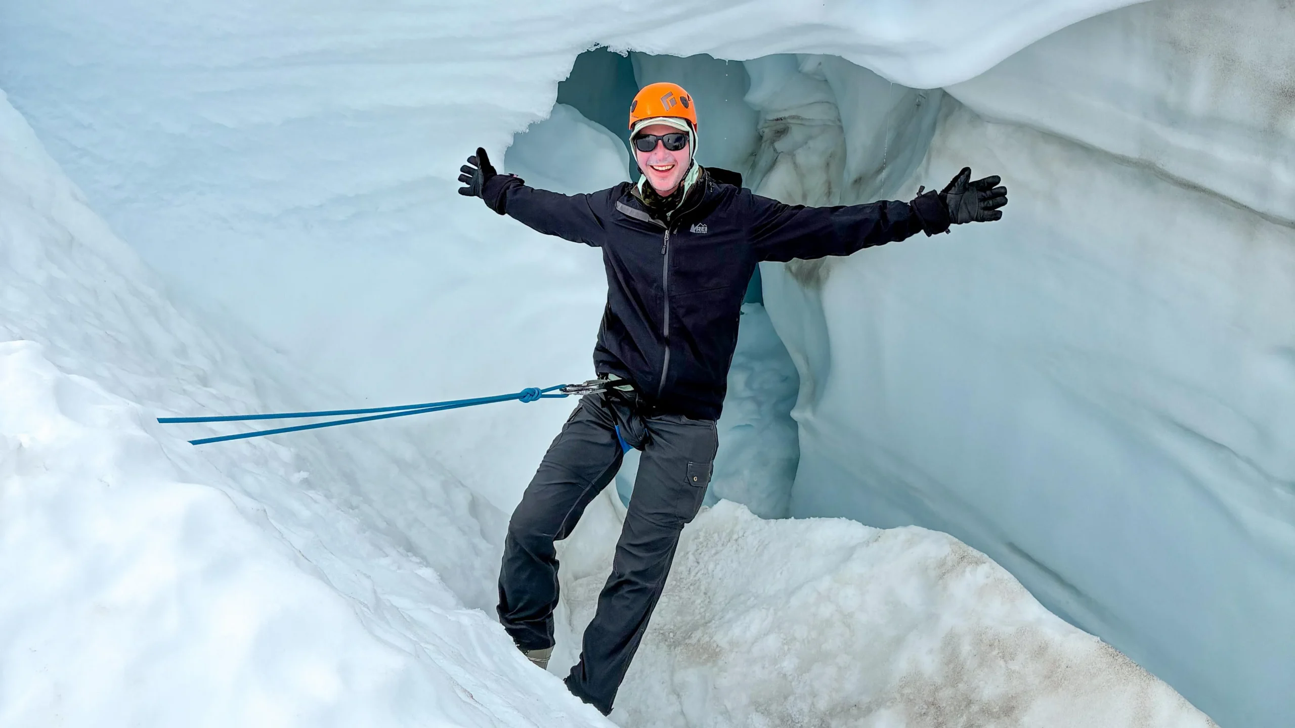 a man in an orange helmet and black winter gear stands on an ice wall while roped into a harness