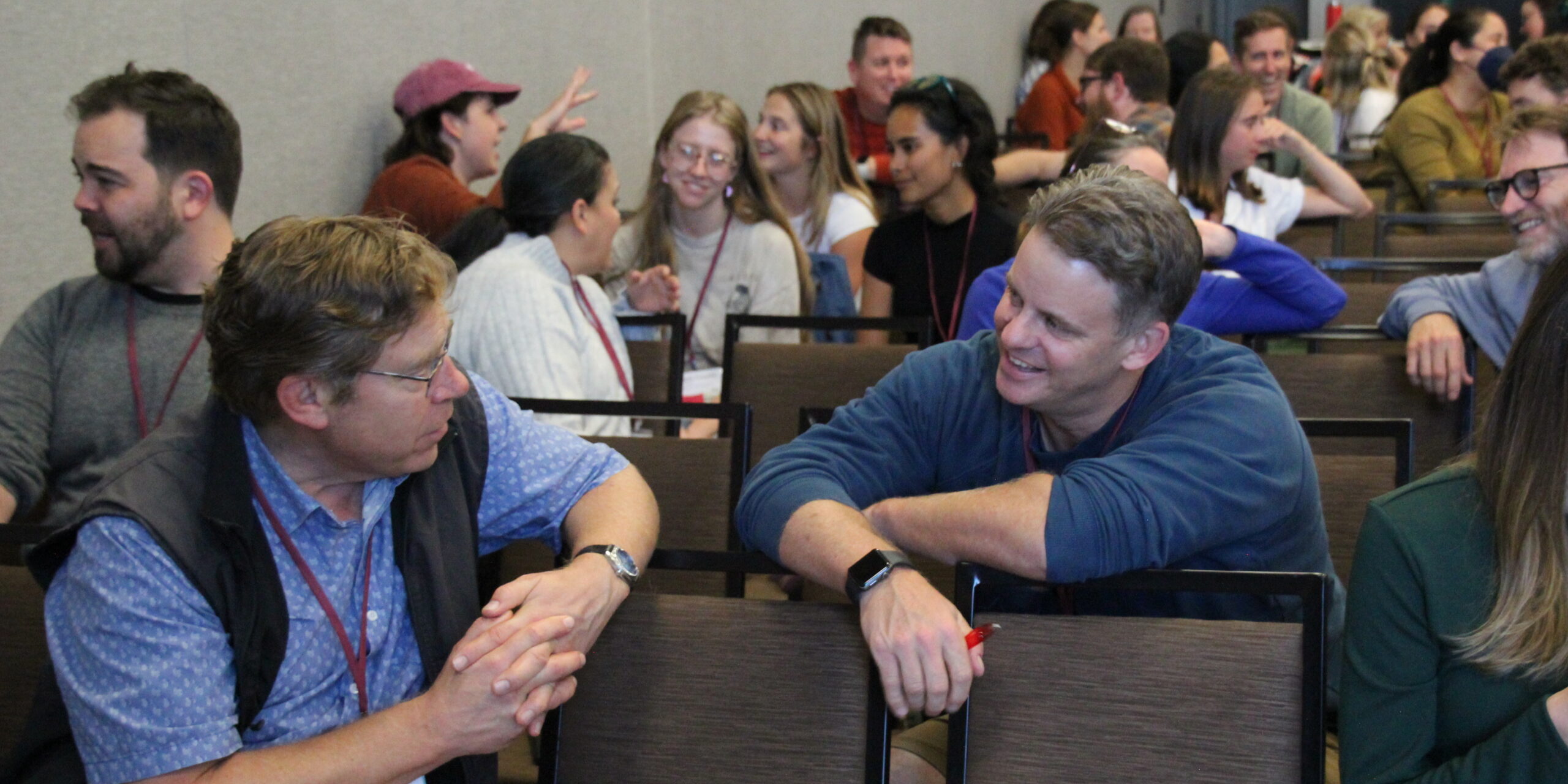 two men in a classroom talk to one another across a chair. many people in the background are engaged in conversation